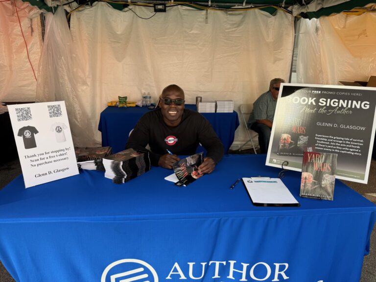Author at book signing event smiling behind a table with promotional materials.
